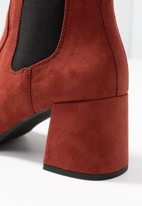 Gros plan sur le talon d'une botte en suède couleur rouille avec un panneau latéral élastique noir et un talon carré sur fond blanc.