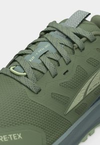 Πράσινο ορειβατικό παπούτσι με κορδόνια με σχέδιο, θηλιά για τράβηγμα, ανθεκτικό επάνω μέρος από διαπνέον πλέγμα και ορατά χαρακτηριστικά "GORE-TEX" και "LONE PEAK 6 GTX".