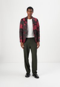 Blazer mit Knopfleiste und rotem Blumenmuster auf schwarzem Stoff, kombiniert mit einem weißen T-Shirt und olivgrünen Hosen, abgerundet mit schwarzen Schuhen.