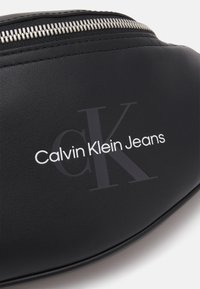 Crna fau kože fanny pack s srebrnim patentnim zatvaračem, utisnutim logotipom i podebljanim bijelim tekstom "Calvin Klein Jeans" na prednjoj strani.