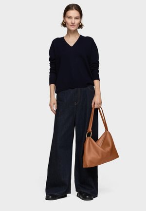 Donna con maglione nero a scollo a V e jeans scuri a gamba larga che tiene una borsa di pelle marrone su uno sfondo grigio uniforme.