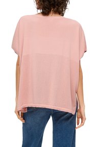 Persona vista da dietro, che indossa un ampio top a maglia rosa con dettagli in rete nella parte bassa della schiena e jeans blu, in piedi su uno sfondo bianco.