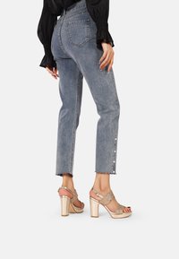 Jeans azzurri chiari a vita alta con gamba dritta e orlo sfilacciato, abbinati a sandali con tacco alto metallici dorati con cinturini intricati.