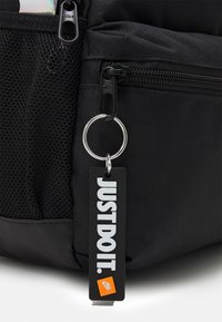 Mochila de tela negra con un bolsillo de malla, con cremalleras negras, un anillo plateado y una etiqueta rectangular con "SIMPLEMENTE HAZLO" y el logo de Nike.