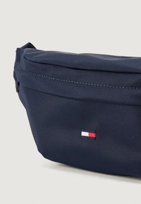 Tamsiai mėlynas audinio fanny pack su kreiva forma. Turi užtrauktuko uždarymą, tekstūruotą paviršių ir mažą logotipą raudonos, baltos ir mėlynos spalvų.