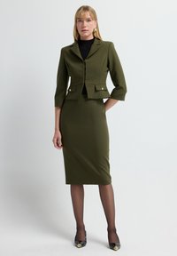 Olivgrünes maßgeschneidertes Blazer mit Dreiviertelärmeln und goldenen Knopf-Accents, kombiniert mit einem taillierten Rock aus demselben Material und in derselben Farbe.