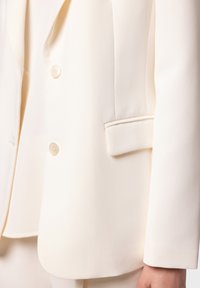 Blazer bianco caratterizzato da una texture liscia, due bottoni e una tasca singola sul lato sinistro, con linee pulite e un design strutturato.
