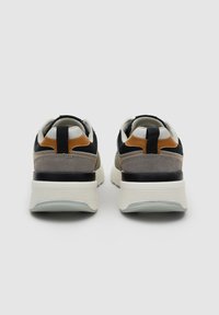 Par de zapatillas deportivas gruesas con suelas blancas, ante gris, malla negra y detalles de cuero marrón, vistas desde atrás sobre un fondo liso.