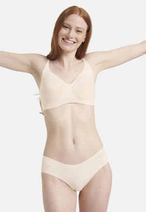 Donna sorridente dai capelli rossi che indossa un reggiseno senza cuciture beige e biancheria intima abbinata, con le braccia sollevate davanti a uno sfondo bianco uniforme.