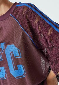 Burgundy mesh top with lace details on one sleeve, blue horizontal stripes, and bold blue graphic numbers on the front. Textured fabric.
