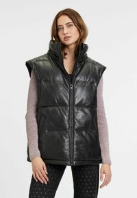 Zwarte leren mouwloze vest met een hoge kraag, rits aan de voorkant en een doorstikte ontwerp. Gecombineerd met een lichtpaarse longsleeve.