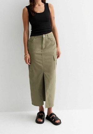 Femme portant un haut noir sans manches, une jupe midi vert olive avec une fente à l'avant et des poches latérales, ainsi que des sandales noires à bout ouvert.