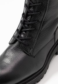 Primo piano di un boot in pelle nera con lacci, dalla superficie testurizzata e suola in gomma spessa, su uno sfondo bianco.