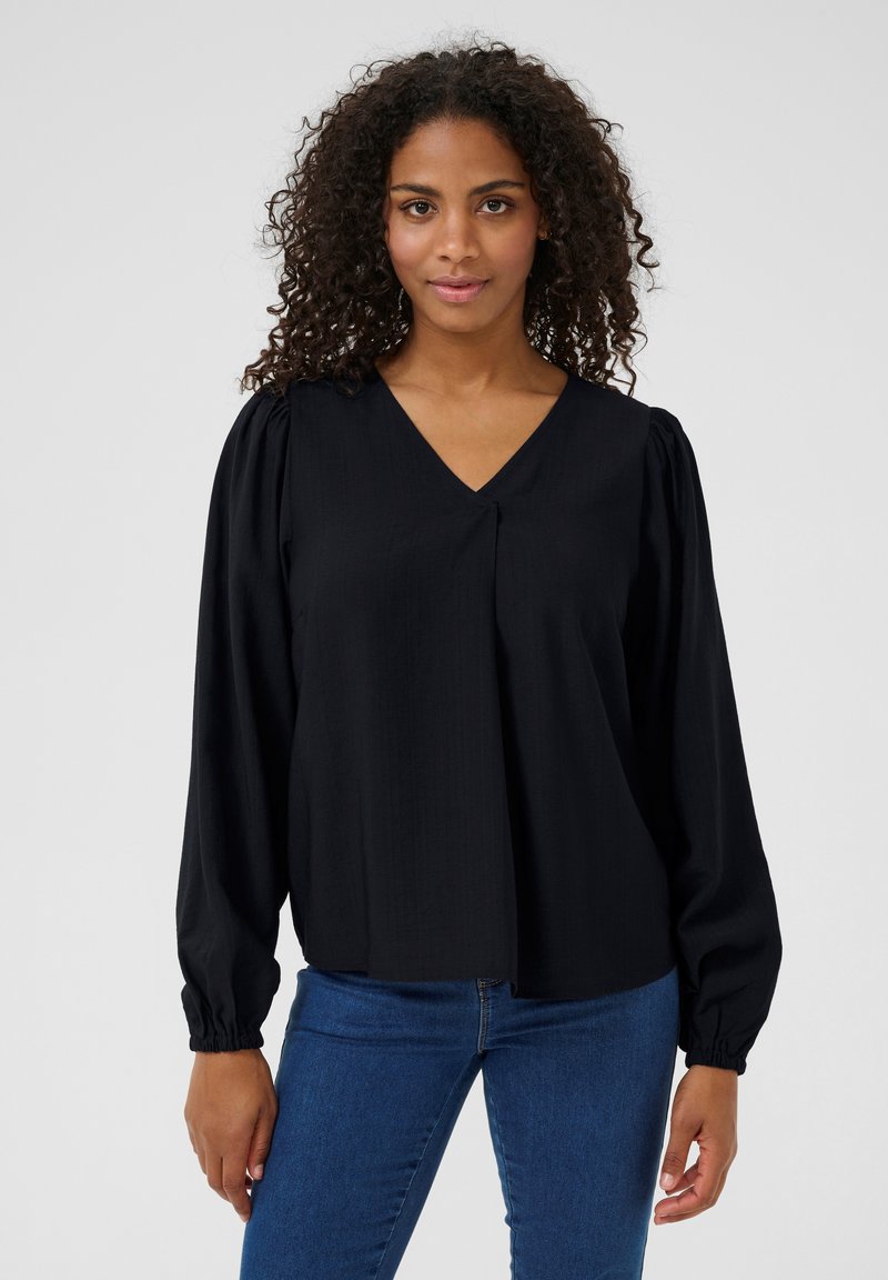 Blouse noire à col en V, avec des manches longues froncées et un détail plissé à l'avant. Le tissu a une texture lisse.