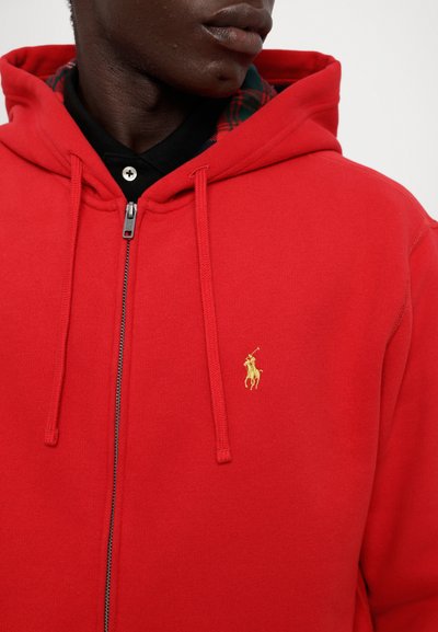 Sudadera roja con cremallera hecha de una mezcla de algodón, con un acento de logo dorado y un forro de cuadros cerca de la capucha. Tela texturizada visible.