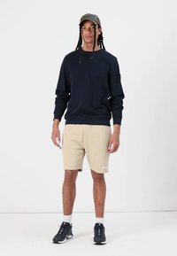 Námornícky sveter, béžové šortky a čierne športové topánky. Model má na sebe maskáčovú čiapku. Hladká tkanina, uvoľnený strih, minimálne značenie.