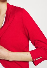 Cárdigan de punto rojo con cuello de chal y mangas de tres cuartos. Presenta una textura suave y botones decorativos en los puños.