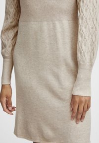 Beiges gestricktes Kleid mit geripptem Rundhalsausschnitt und langen, Kabelstrickärmeln. Glatte Textur, figurbetonte Silhouette, fällt oberhalb des Knies.