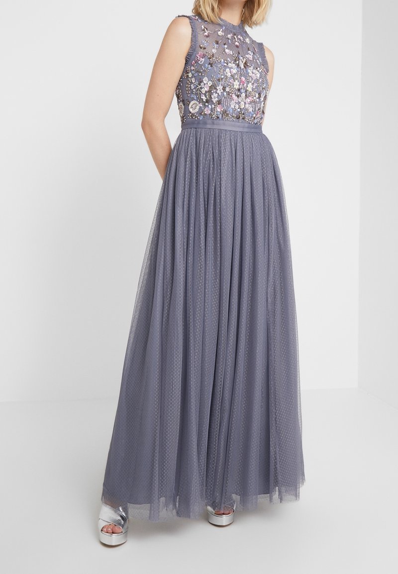 Femme portant une robe en tulle gris sans manches avec une broderie florale sur le corsage et des sandales à plateforme argentées, debout devant un fond uni.