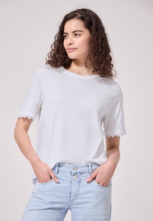 Femme aux cheveux bouclés portant une chemise blanche à manches courtes avec un bord en dentelle et un jean bleu clair, mains dans les poches, regardant sur le côté.
