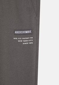 Pantalon de survêtement gris avec une étiquette noire cousue portant "ABERCROMBIE" et un texte blanc en dessous, fabriqué à partir d'un tissu doux avec une texture lisse.