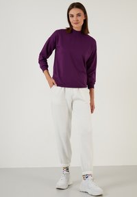 Felpa viola con collo alto, maniche lunghe e polsini elasticizzati, abbinata a pantaloni jogger bianchi e sneakers bianche.