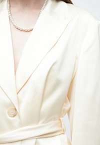 Kreminės spalvos švarkas su šalio tipo apykakle, vienu užsegamu mygtuku ir dirželiu liemenyje. Lygios tekstūros, pritaikymo liemeniui ir struktūruotais pečiais.