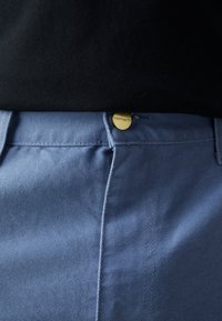 Lichtblauwe broek van gestructureerde stof met een gouden knoopsluiting, zicht op de tailleband en stikdetails. Zwarte blouse erboven.