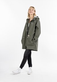 Pelerină de ploaie impermeabilă, verde, cu glugă, două buzunare în față, accesorii argintii și șnururi ajustabile, purtată cu blugi negri și adidași albi.