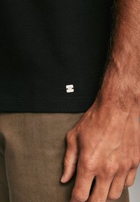 Polo-shirt noir texturé avec une étiquette logo argentée à l'ourlet ; porté avec un pantalon marron clair, montrant une main reposant contre le tissu.