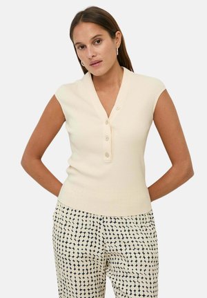 Mujer que lleva un top sin mangas color crema con botones y pantalones blancos de talle alto con un patrón de puntos geométricos negros, de pie con las manos detrás de la espalda.