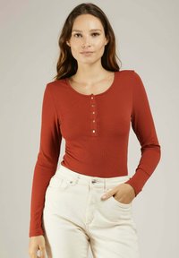 Haut à manches longues de couleur rouille en texture côtelée avec un col rond et une patte de boutonnage à cinq boutons, associé à un pantalon blanc taille haute.
