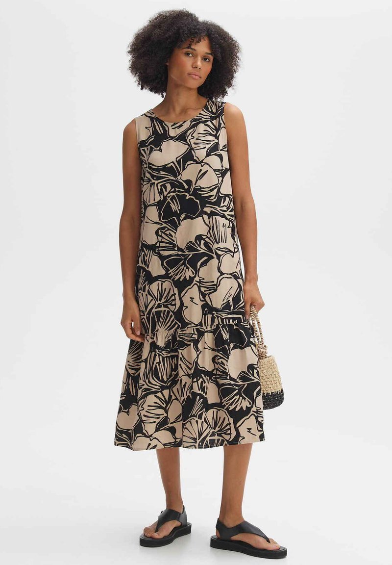 Floral-patterned sleeveless dress in beige and black, featuring a relaxed fit and tiered hem. Worn with black sandals and a woven bag.