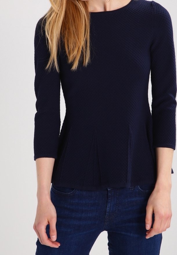 Femme portant un haut texturé bleu marine à manches longues avec une taille cintrée et un jean bleu foncé, debout devant un fond uni.