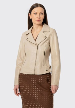 Donna che indossa una giacca di pelle beige con tasche con cerniera sopra una gonna marrone con pois bianchi, in piedi contro uno sfondo semplice.