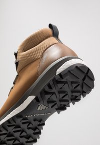 Botte de randonnée marron avec une tige texturée, une semelle en caoutchouc et des crampons profonds. Comprend un système de laçage et une languette pour faciliter l'enfilage.