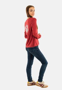 Rode lange mouwen shirt met een grafisch ontwerp op de achterkant, gecombineerd met donkerblauwe skinny jeans en beige sneakers met gele en bordeauxrode strepen.