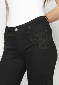 Black jeans featuring floral embroidery in silver thread, five-pocket design, metal button closure, and slight stretch fabric for comfort.