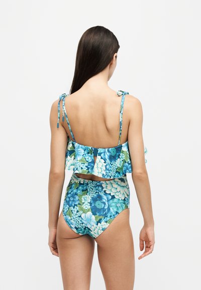 Femme aux longs cheveux foncés portant un maillot de bain deux pièces bleu à motifs floraux avec un haut volanté et un bas taille haute, vue de dos sur un fond uni.
