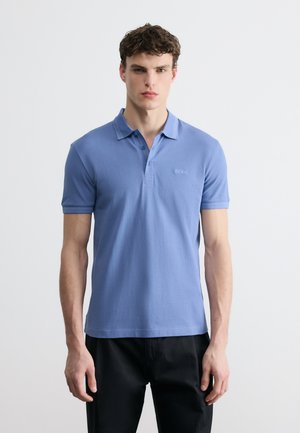 Νεαρός άνδρας που φοράει ανοιχτό μπλε κοντομάνικο πόλο με γιακά και μαύρο παντελόνι, στέκεται μπροστά σε έναν απλό λευκό φόντο.