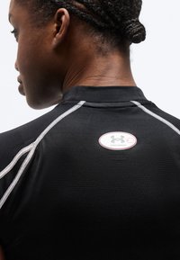 Άτομο με πλεξούδες που φοράει μαύρο αθλητικό μπλουζάκι με λευκές ραφές και λογότυπο Under Armour στο πάνω μέρος της πλάτης.