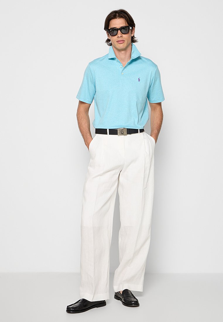 Jasnoniebieska koszulka polo z krótkim rękawem i kołnierzykiem, zestawiona z luźnymi białymi spodniami i czarnymi butami eleganckimi. Noszone czarne okulary przeciwsłoneczne.