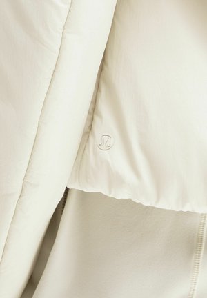 Približnji posnetek kremasto obarvane podložene jakne z vidnim šivom in majhnim logotipom Lululemon na površini tkanine.