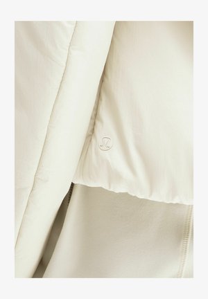 Približnji posnetek kremasto obarvane podložene jakne z vidnim šivom in majhnim logotipom Lululemon na površini tkanine.