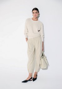 Femme debout en pull crème à texture pointillée, pantalon plissé beige, ballerines noires à bouts pointus et tenant un grand sac à main crème.