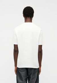 Άνδρας που φοράει απλό λευκό κοντομάνικο μπλουζάκι και σκούρο τζιν, στέκεται γυρισμένος με την πλάτη σε ένα απλό ανοιχτό γκρι φόντο.