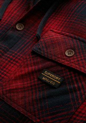Camisa de franela a cuadros en rojo y negro con dos botones marrones, un bolsillo en el pecho y una etiqueta negra que dice "SUPERDRY MFG/CO" cosida en amarillo.