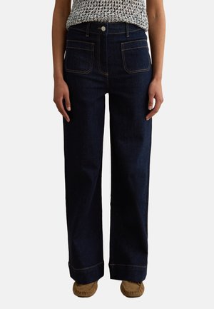 Femme portant un jean large bleu foncé avec des poches plaquées à l'avant, des ourlets retroussés, un bouton argenté, associée à des mocassins marron.