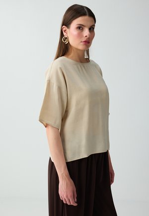 Frau mit langen braunen Haaren trägt ein beiges Kurzarmtop, große goldene Spiralohrringe und dunkelbraune Hose, steht vor hellem Hintergrund.