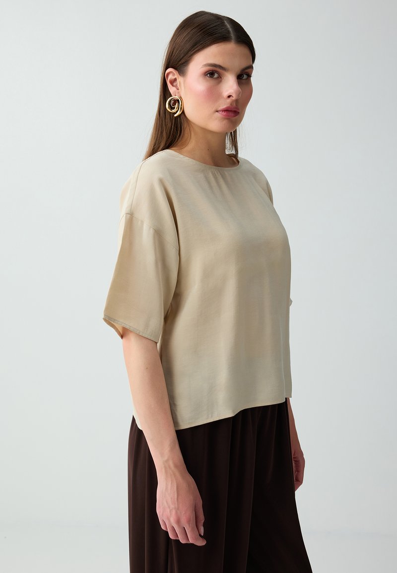 Femme aux longs cheveux bruns porte un haut beige à manches courtes, de grandes boucles d'oreilles dorées en spirale et un pantalon brun foncé, debout devant un fond clair.